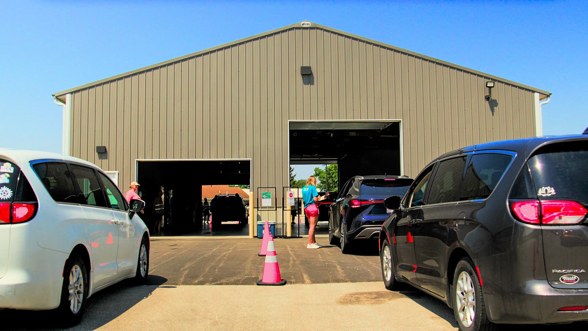 Minivans lined up outside a drive-thru meal pickup site as a volunteer checks in a family at a 2026 summer distribution